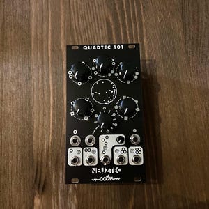 Puede incluir: Módulo Eurorack Quadtec 101 negro con botones y conectores. El módulo tiene una etiqueta blanca con el texto "Quadtec 101" y "~cct~" en una fuente estilizada.