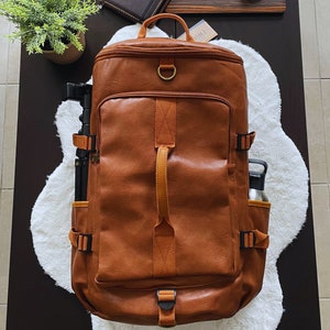 May include: A brown leather backpack with orange straps and buckles. The backpack has a zipped front pocket and a side pocket for a water bottle. The backpack is sitting on a white fluffy rug.