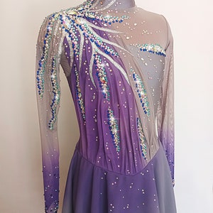Op de afbeelding: Een paars-grijze kunstschaatsjurk met lange mouwen en een hoge hals. De jurk is versierd met strass en pailletten in verschillende kleuren, waaronder blauw, zilver en groen.