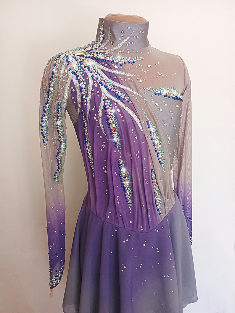 Op de afbeelding: Een paars-grijze kunstschaatsjurk met lange mouwen en een hoge hals. De jurk is versierd met strass en pailletten in verschillende kleuren, waaronder blauw, zilver en groen.