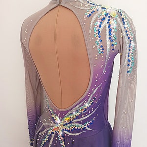 Op de afbeelding: Een langarmige, paars- en grijze ombre schaatsjurk met een grote, open rug. De jurk is versierd met zilveren en iriserende strass steentjes in een bloemmotief.
