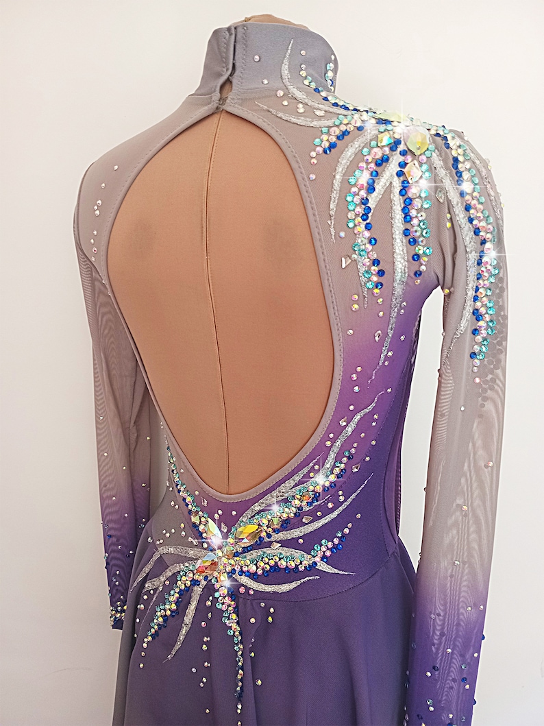 Op de afbeelding: Een langarmige, paars- en grijze ombre schaatsjurk met een grote, open rug. De jurk is versierd met zilveren en iriserende strass steentjes in een bloemmotief.