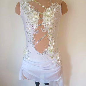 May include: A white figure skating dress with floral and rhinestone embellishments. The sleeveless dress features a high neckline, sheer mesh panels, and a draped skirt. The bodice is adorned with white flowers and sparkling rhinestones.