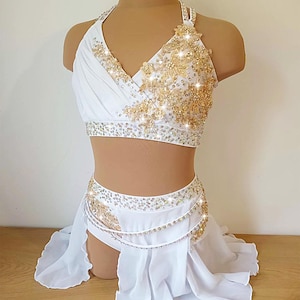 May include: White dance costume with gold detailing. The halter top and skirt set features gold floral designs and sparkling accents. The skirt has a flowing, layered design, creating an elegant look. The costume is displayed on a mannequin.