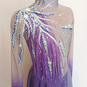 Op de afbeelding: Een paars en grijs kunstschaatsjurk met lange mouwen en een hoge hals. De jurk is versierd met strass en pailletten in verschillende kleuren, waaronder blauw, groen en zilver.
