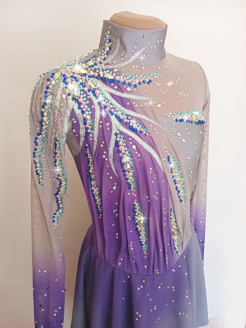 Op de afbeelding: Een paars en grijs kunstschaatsjurk met lange mouwen en een hoge hals. De jurk is versierd met strass en pailletten in verschillende kleuren, waaronder blauw, groen en zilver.