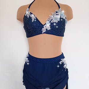 May include: A navy blue dance costume set. The halter top is embellished with sparkling beads and floral accents. The matching skirt has a draped design and is also adorned with floral details. The costume is displayed on a mannequin.