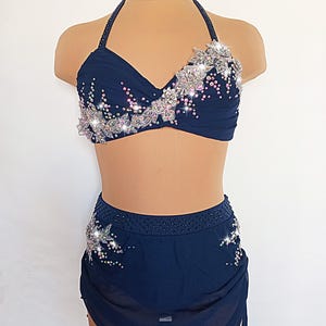 Könnte beinhalten: Ein marineblaues Tanzkostüm-Set. Das Neckholder-Top und der Rock sind mit silbernen und irisierenden Strasssteinen verziert. Das Top hat florale Designs, und der Rock hat ein fließendes, asymmetrisches Design.