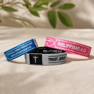 Pode incluir: Três pulseiras elásticas em azul, rosa e cinza claro. A pulseira azul apresenta o texto "I CAN DO ALL THINGS THROUGH CHRIST WHICH STRENGTHENETH ME". A pulseira rosa exibe "PHILIPPIANS 4:13". A pulseira cinza mostra uma cruz e "TRUST JESUS".