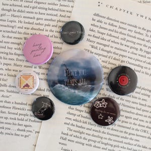 Puede incluir: Una colección de botones redondos con varios diseños y texto. Un botón dice "I LOVE YOU. THAT'S ALL" sobre una imagen de mar tormentoso. Otros botones tienen frases como "hang painting here!" y gráficos de estrellas.