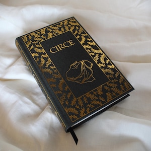 Op de afbeelding: Een hardcover boek getiteld "CIRCE" met een zwarte kaft en gouden blad details. De kaft heeft gouden tekst en een gouden illustratie. Een zwart lint-bladwijzer is zichtbaar. Het boek ligt op een witte stoffen ondergrond.