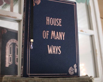 May include: A navy blue hardback book with the title "HOUSE OF MANY WAYS" in rose gold lettering. The book has a black ribbon bookmark with a gold charm and a purple crystal.
