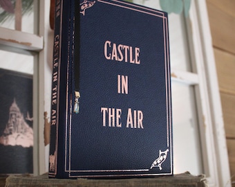 May include: A dark blue leather-bound journal with a rose gold title that reads "CASTLE IN THE AIR".