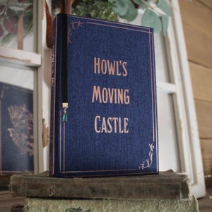 May include: A blue book with a black ribbon bookmark and a green charm. The book has the title "Howl's Moving Castle" printed in gold lettering.
