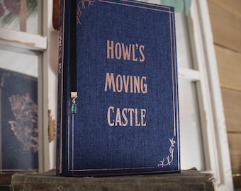 May include: A blue book with a black ribbon bookmark and a green charm. The book has the title "Howl's Moving Castle" printed in gold lettering.