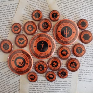 Pode incluir: Uma coleção de pins circulares laranja e pretos, cada um com o nome e o símbolo de um deus ou deusa grega, com uma borda decorativa. Os pins estão dispostos sobre um fundo de páginas de livro abertas.