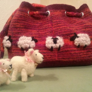 Könnte beinhalten: Eine rot-orange gestreifte Stricktasche mit Kordelzug und Schaf-Applikationen. Im Vordergrund befinden sich zwei weiße, flauschige Schafspielzeuge.