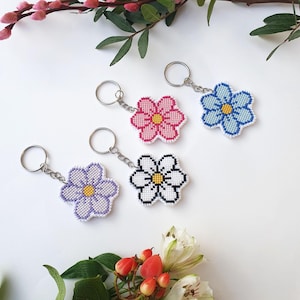 以下が含まれることがあります： 異なる色の花が付いた花の形をしたキーホルダーが4つ。キーホルダーは白い布でできており、ピンク、青、紫、白の花が刺繍されています。