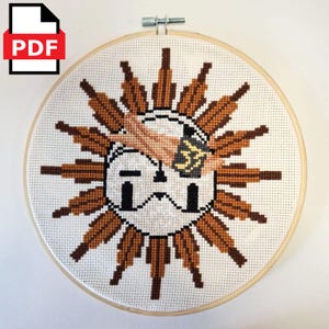 Può includere: Un disegno a punto croce di un sole con raggi marroni e un volto, che tiene una tazza di caffè. Il disegno è in un telaio da ricamo in legno. Un'icona PDF si trova nell'angolo in alto a sinistra.