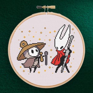 May include: A cross-stitch embroidery hoop featuring two pixelated characters from a video game. One character wears a brown hat and cloak, the other a red cape. The background is light pink with yellow star accents, set in a wooden hoop.