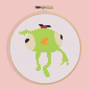 May include: Cross-stitch art of a green creature with large eyes and an orange belly, wearing a small hat. The design is set in a wooden embroidery hoop against a pink background. The creature has a whimsical, cartoonish appearance.