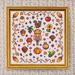 May include: Cross-stitch artwork in a gold frame featuring autumn-themed elements. The design includes a scarecrow, pumpkins, corn, acorns, a sunflower, and other fall harvest symbols. The artwork is set against a white background.