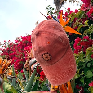 Puede incluir: Una gorra de béisbol de color terracota con un pequeño diseño de flor bordado en la parte delantera. La gorra está sentada en una rama de una planta de ave del paraíso con flores naranjas y amarillas.