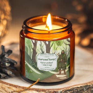Puede incluir: Una vela encendida en un frasco de vidrio marrón con una etiqueta que presenta una escena de bosque y el texto "Sherwood Forest Mythic Woodland Cedar & Sage". La vela está sobre una superficie de madera.