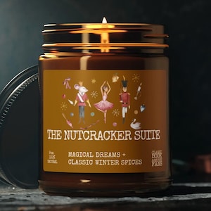 Può includere: Un barattolo di vetro marrone per candele con un coperchio dorato. L'etichetta presenta un'illustrazione fantasiosa di personaggi del balletto Lo Schiaccianoci, tra cui un re topo, una ballerina e un soldato schiaccianoci. Il testo sull'etichetta recita "The Nutcracker Suite" e "Magical Dreams + Classic Winter Spices".