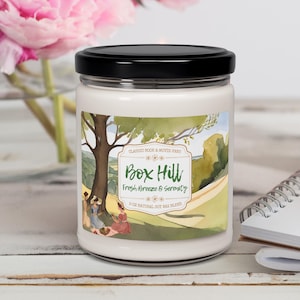 May include: A white candle jar with a black lid. The label features a watercolor illustration of a green hillside with a tree and people picnicking. The text on the label reads "CLASSIC BOOK & MOVIE FANS Box Hill Fresh Breeze & Serenity 9 OZ NATURAL SOY WAX BLEND".