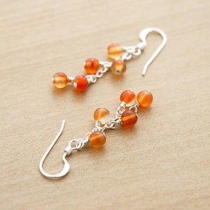 May include: A pair of dangle earrings featuring small, round, orange and amber-colored beads. The beads are strung together with silver-colored chain and have curved ear wires.