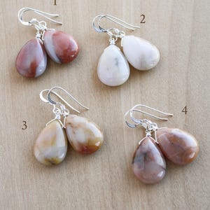 May include: Four pairs of teardrop-shaped gemstone earrings with silver-colored hooks. The gemstones vary in color, including shades of red, white, and brown. The earrings are displayed on a light brown wooden surface.
