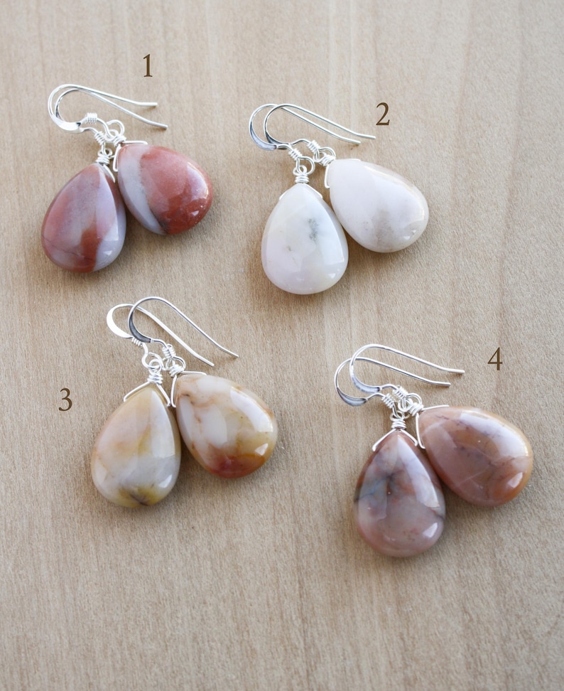 May include: Four pairs of teardrop-shaped gemstone earrings with silver-colored hooks. The gemstones vary in color, including shades of red, white, and brown. The earrings are displayed on a light brown wooden surface.