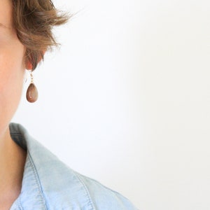 May include: A close-up of a brown teardrop-shaped earring with a gold-colored hook. The earring is hanging from an ear. The background is white.