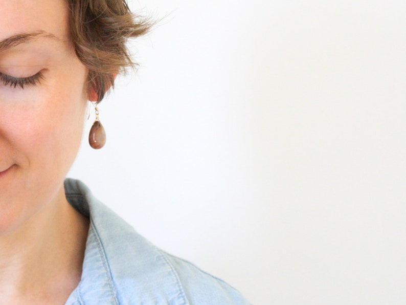 May include: A close-up of a brown teardrop-shaped earring with a gold-colored hook. The earring is hanging from an ear. The background is white.