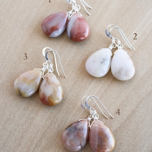 May include: Four pairs of gemstone earrings with silver-colored ear wires. The gemstones are teardrop-shaped and vary in color, including shades of pink, white, and brown. The earrings are displayed on a light brown surface.