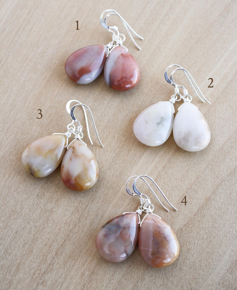 May include: Four pairs of gemstone earrings with silver-colored ear wires. The gemstones are teardrop-shaped and vary in color, including shades of pink, white, and brown. The earrings are displayed on a light brown surface.