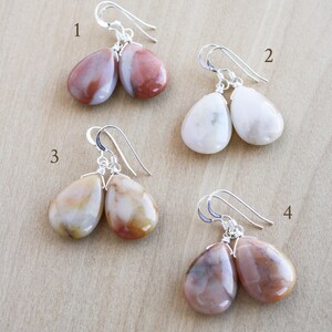 May include: Four pairs of gemstone earrings with silver-colored ear wires. The earrings feature teardrop-shaped stones in various colors, including white, red-brown, and cream. The earrings are displayed on a light brown surface.