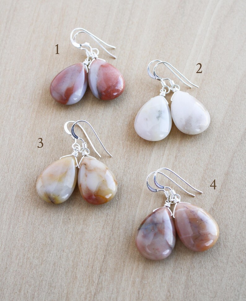 May include: Four pairs of gemstone earrings with silver-colored ear wires. The earrings feature teardrop-shaped stones in various colors, including white, red-brown, and cream. The earrings are displayed on a light brown surface.
