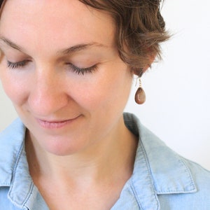 May include: A close-up of a person wearing a pair of teardrop-shaped earrings. The earrings are a warm brown color and hang from gold-colored hooks. The person is wearing a light blue denim shirt.
