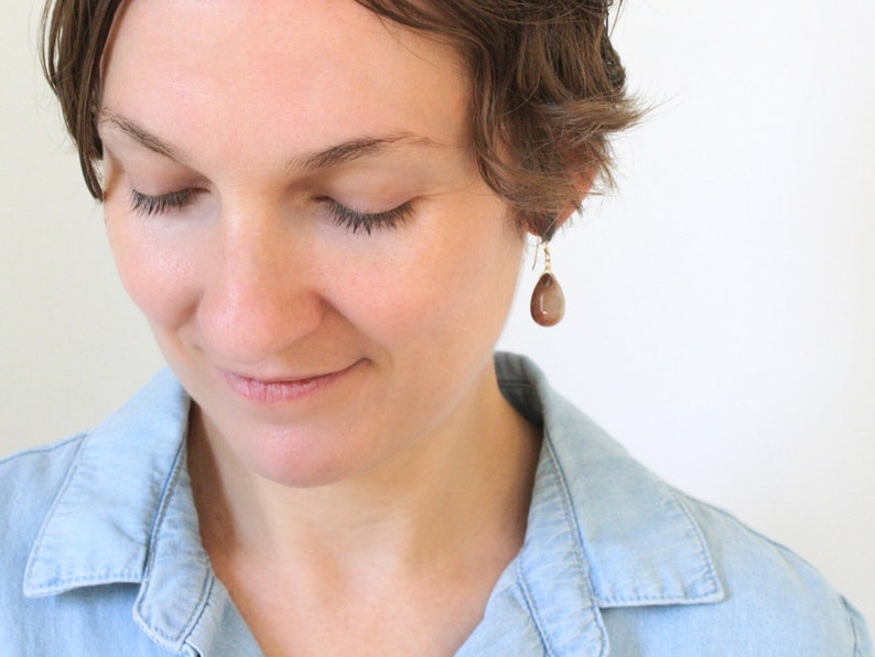 May include: A close-up of a person wearing a pair of teardrop-shaped earrings. The earrings are a warm brown color and hang from gold-colored hooks. The person is wearing a light blue denim shirt.