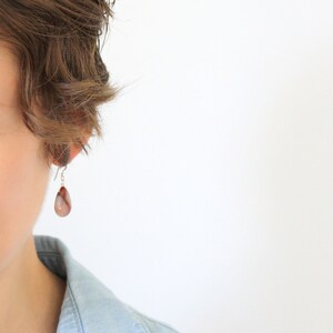 May include: A close-up of a person wearing a single teardrop-shaped earring. The earring is a light brown color with darker brown and white marbling. The earring hangs from a silver-colored hook.