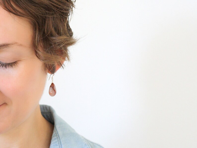 May include: A close-up of a person wearing a single teardrop-shaped earring. The earring is a light brown color with darker brown and white marbling. The earring hangs from a silver-colored hook.