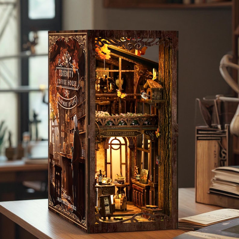 May include: A detailed miniature wooden model of a Serenity Coffee House, featuring the text "Serenity Coffee House" and "Enjoyment of Life" on its facade. The interior is lit, revealing a charming cafe setting with tables, chairs, and decorative features.
