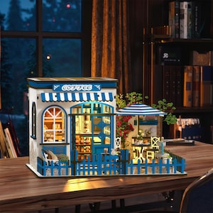 May include: A miniature coffee shop model with a blue and white color scheme. The model features a sign that says "COFFEE", a striped awning, and a small outdoor seating area with a blue and white umbrella. The model is on a wooden table.