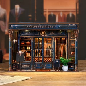 May include: A miniature model of a men's clothing store with a blue and gold facade. The sign reads "GOLDEN SECTION LINE" and "PERSONAL SERVICE". Inside, suits and accessories are displayed. A small chalkboard sign and potted plant are in front.