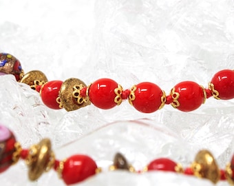 May include: A red and gold beaded necklace with small, round beads and decorative gold spacers. The necklace is lying on a white, textured surface.