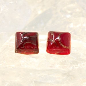 Peut inclure: Une paire de boucles d'oreilles carrées avec une base en verre rouge et des paillettes dorées. Les boucles d'oreilles ont un motif unique et tourbillonnant.