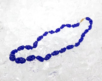 Può includere: Una collana di perle di vetro blu con una chiusura dorata. La collana è adagiata su un letto di ghiaccio bianco.