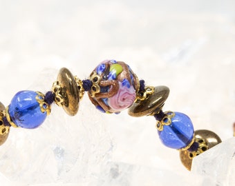 May include: A close-up of a blue and gold beaded bracelet with a pink and gold floral bead in the center.